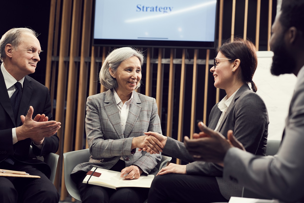 Smiling mature Asian businesswoman with short hair sitting with open diary on knees and handshaking with business networking group leader